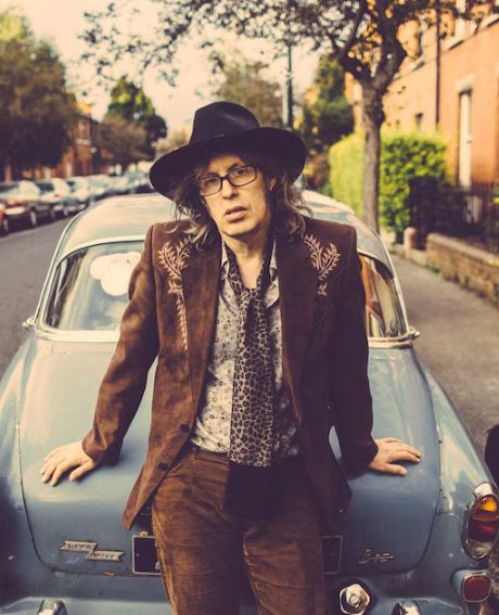 Mike Scott pose adossé à une voiture vintage sur une rue bordée de maisons en briques. Il porte un ensemble en velours marron, composé d'une veste brodée et d'un pantalon assorti,