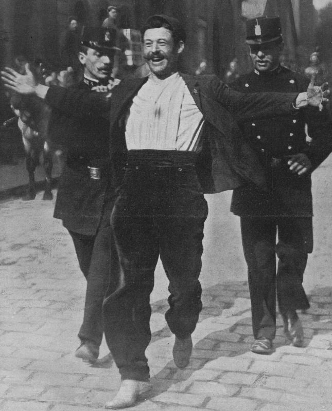 Un homme souriant, vêtu d'une chemise en velours clair et d'une veste sombre, les bras grands ouverts, escorté par deux policiers en uniforme sur une rue pavée.